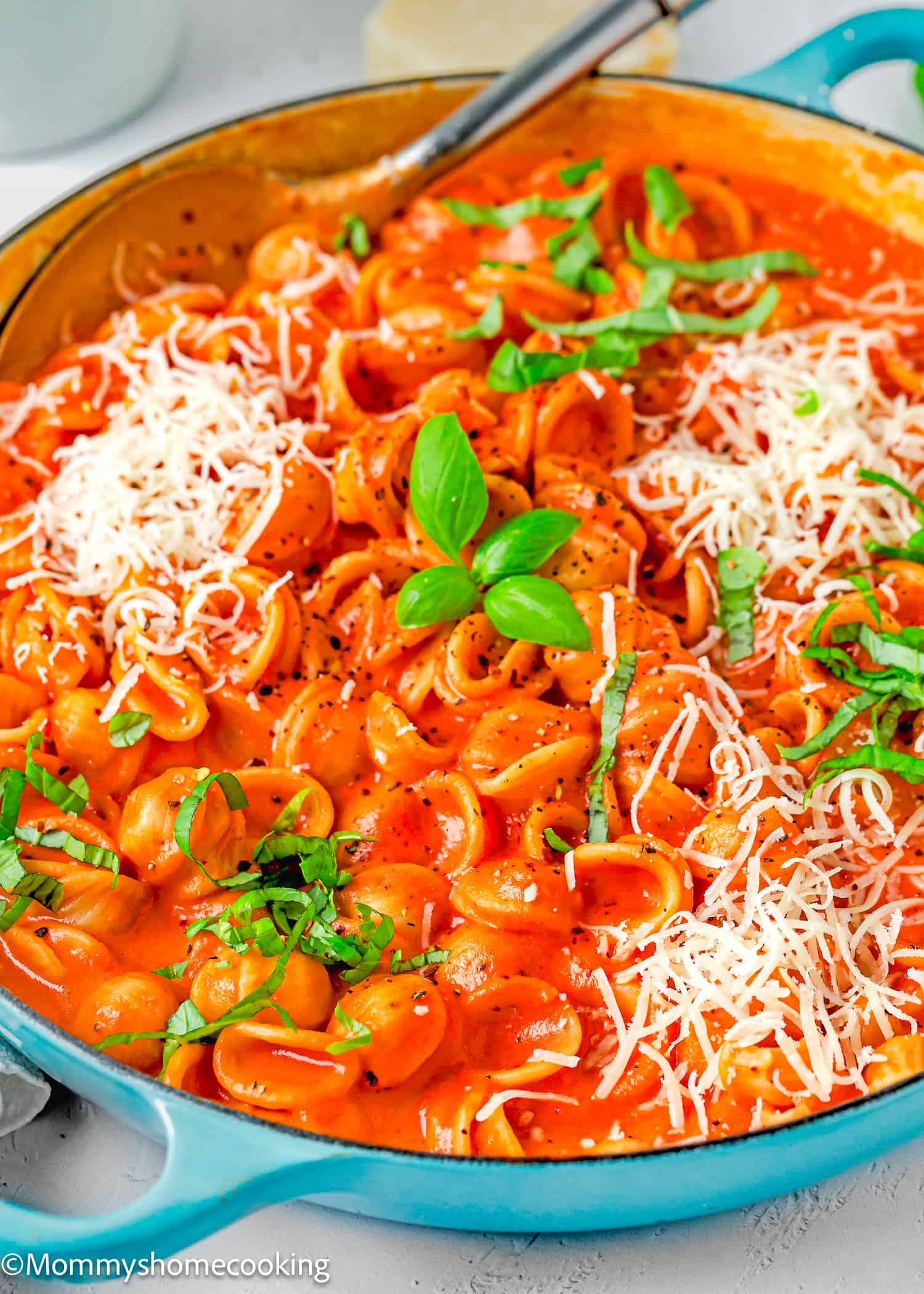 Creamy Tomato Pasta in a blue skillet with a serving spoon, grated cheese and basil over a white surface.
