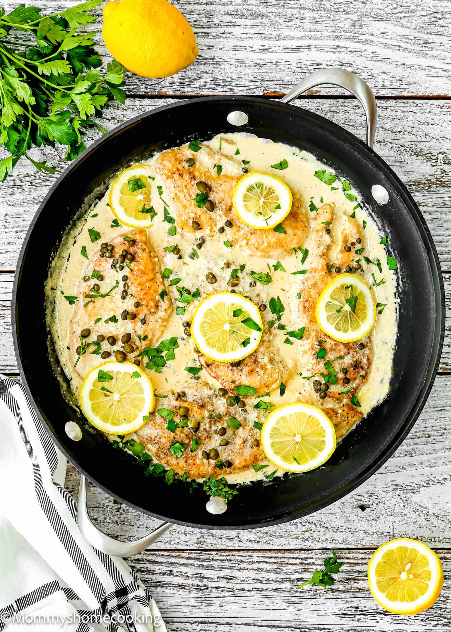Easy Chicken Piccata in creamy sauce in a skilled over a wooden surface with parsley, lemons and kitchen towel on the side.