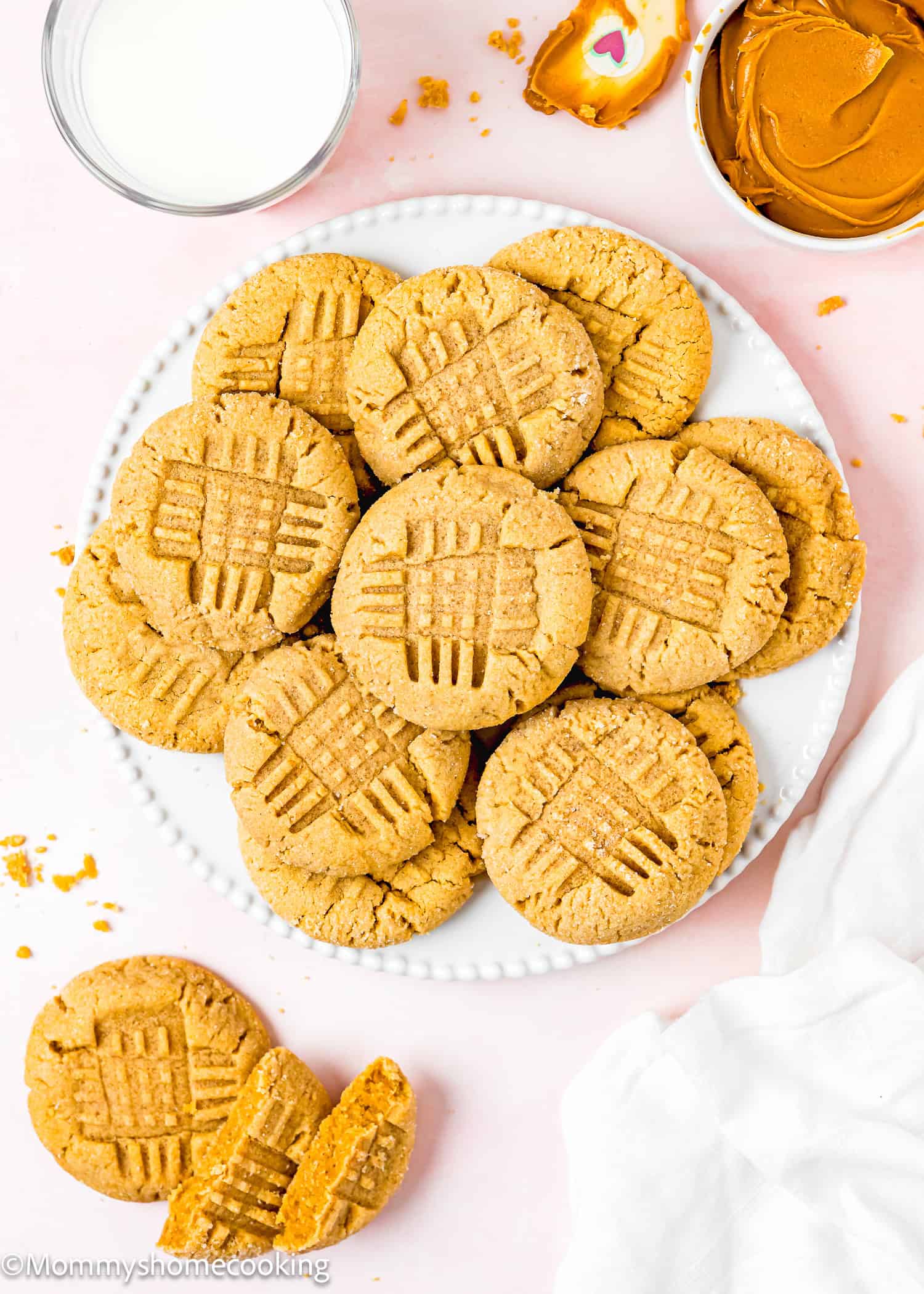 Eggless Peanut Butter Cookies in a a plate over a pink surface with peanut butter, spatula and a glass of milk on the side.