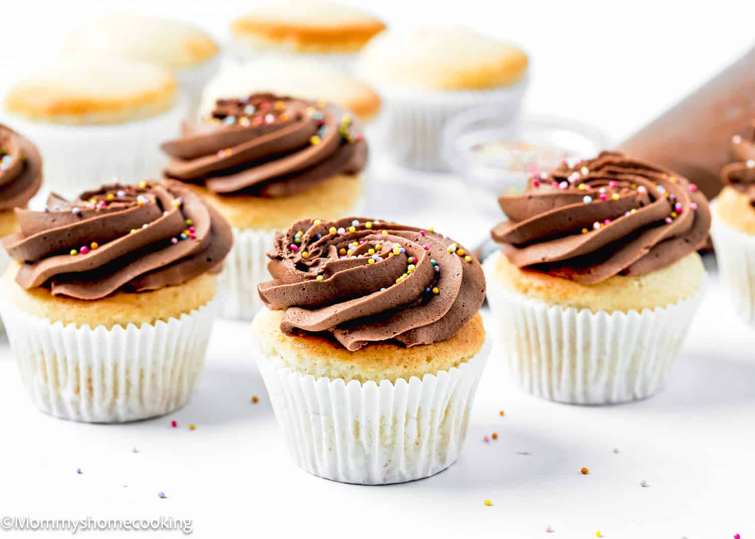 vegan Vanilla Cupcakes with chocolate frosting and sprinkles over a white surface.