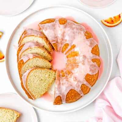 a sliced Simple Orange Bundt Cake without eggs with glaze on a serving plate with a plate and a bowl with orange glaze on the side.