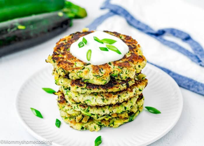 A stack of Easy Crispy Zucchini Fritters (Egg-Free) topped with sour cream and chopped green onions on a white plate, with zucchinis and a white cloth in the background.
