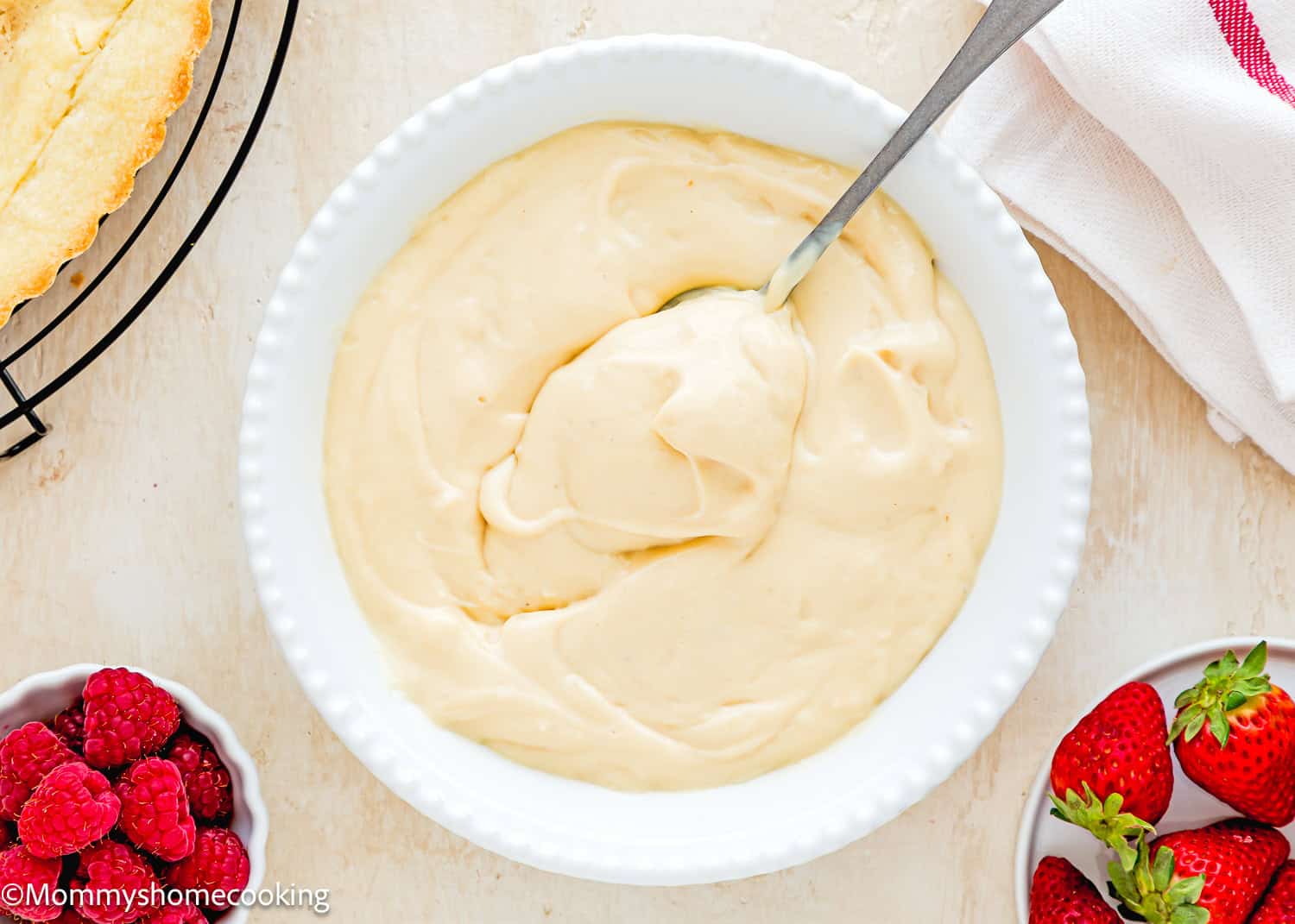 Vegan Pastry Cream in a bowl with a spoon over a beige surface with berries and a tart on the sides.