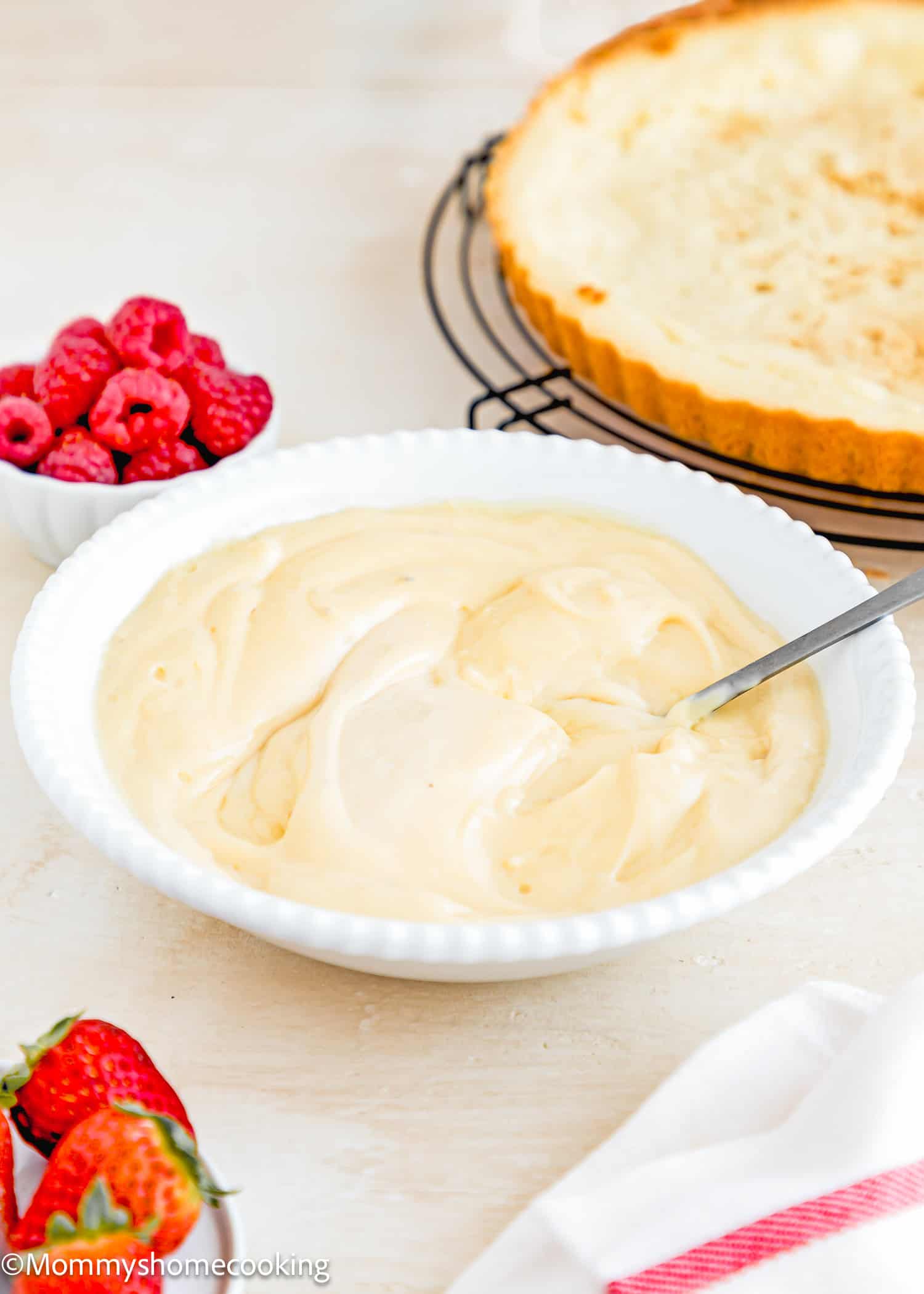 Vegan Pastry Cream in a bowl with a spoon over a beige surface with berries and a tart in the background.