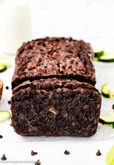Eggless Double Chocolate Zucchini Bread over a piece of parchment paper with fresh zucchini slices and chocolate chips around it and a bottle of milk in the background.