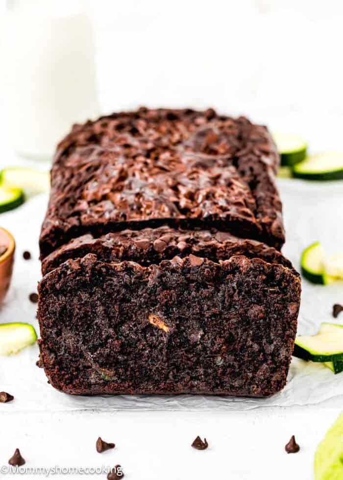 Eggless Double Chocolate Zucchini Bread over a piece of parchment paper with fresh zucchini slices and chocolate chips around it and a bottle of milk in the background.