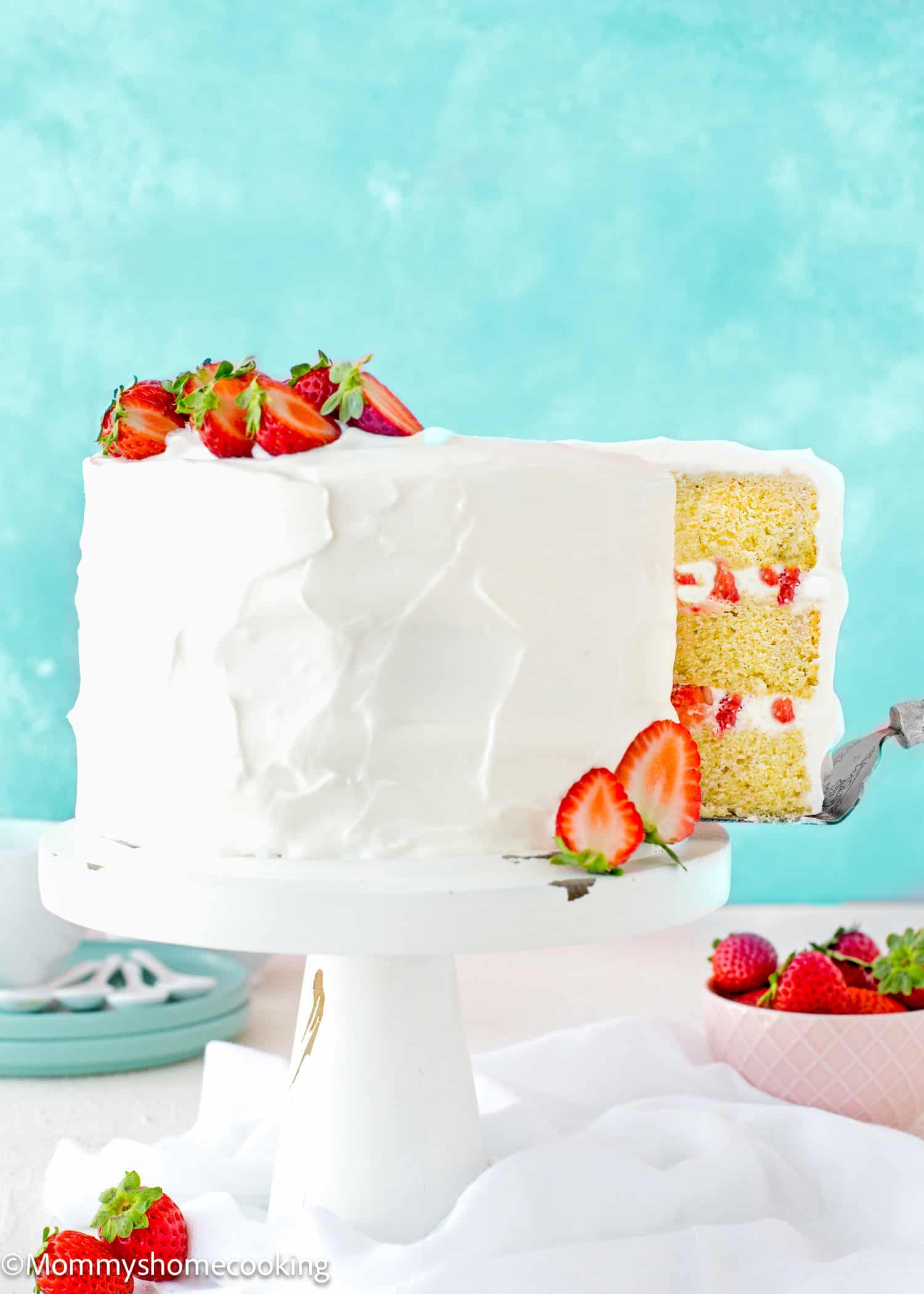 a cake server picking a slice of a Eggless Strawberry Shortcake Cake on a cake stand over a white surface.