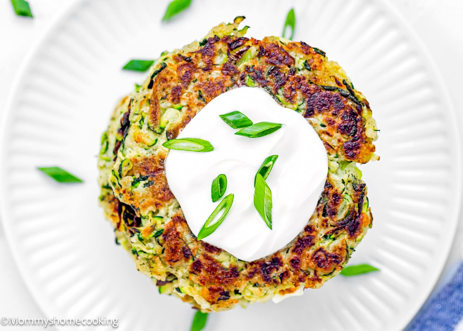 a stack of Eggless Zucchini Fritters on a plate with sour cream on top and chopped green onions.