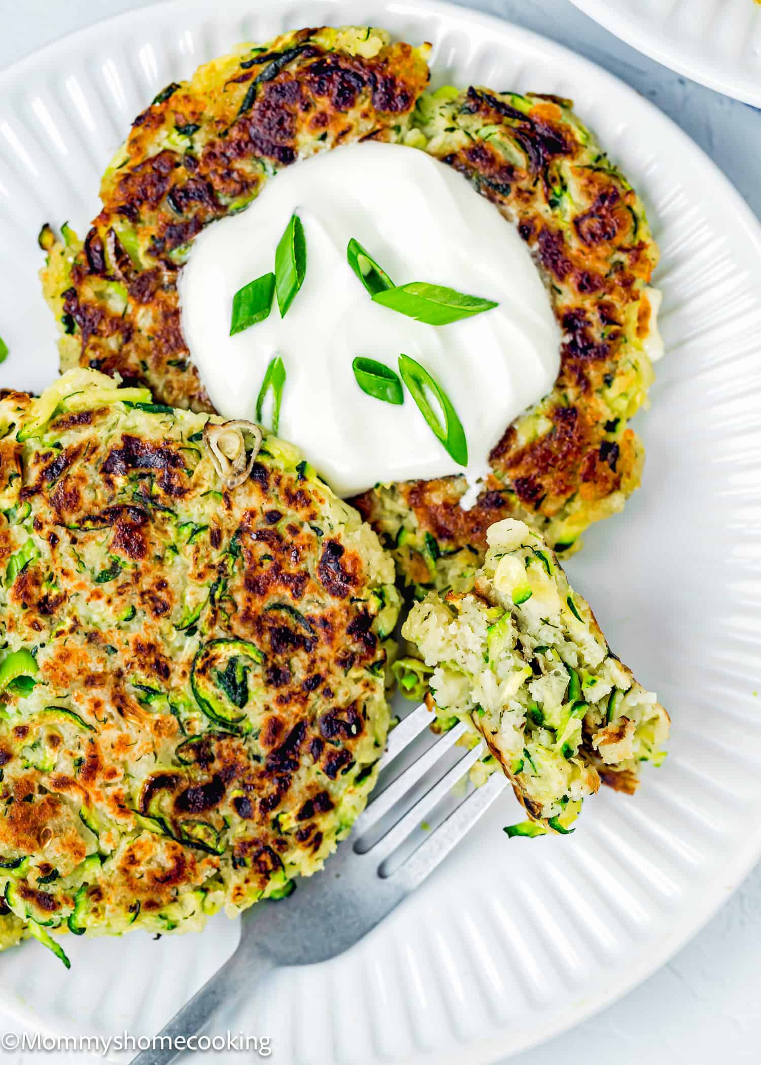 a fork holding a Eggless Zucchini Fritter piece on a plate with two more Eggless Zucchini Fritters.