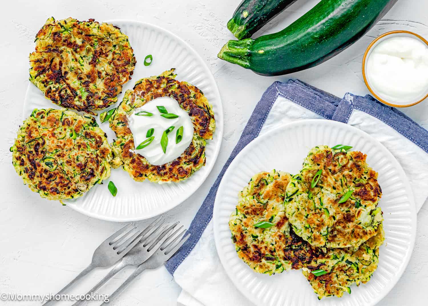 two plates with Eggless Zucchini Fritters over a white surface with a kitchen towel, forks, fresh zucchinis and a bowl with sour cream.