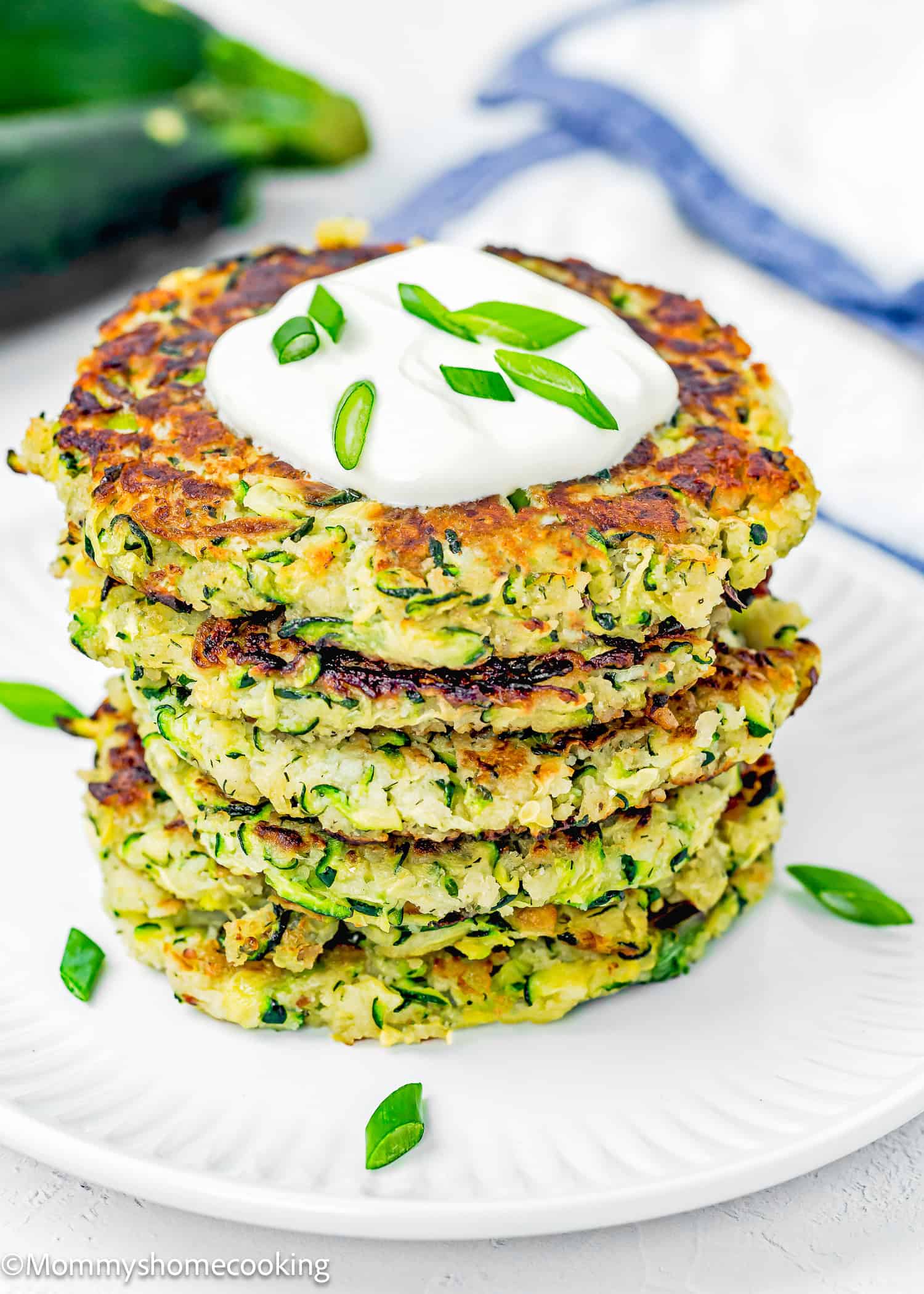 stack of five Eggless Zucchini Fritters over a white plate with sour cream and chopped green onions on top.