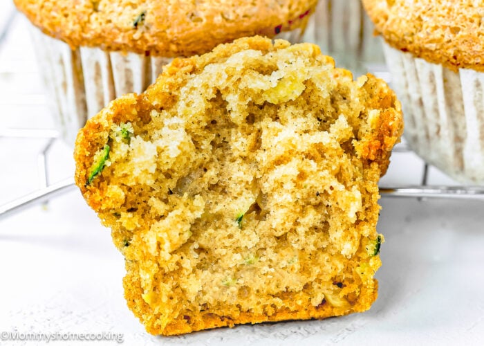 A sliced Eggless Zucchini Muffin (Soft, Moist & Dairy-Free) reveals a tender, crumbly interior, set against a background of whole muffins in white paper liners on a cooling rack.