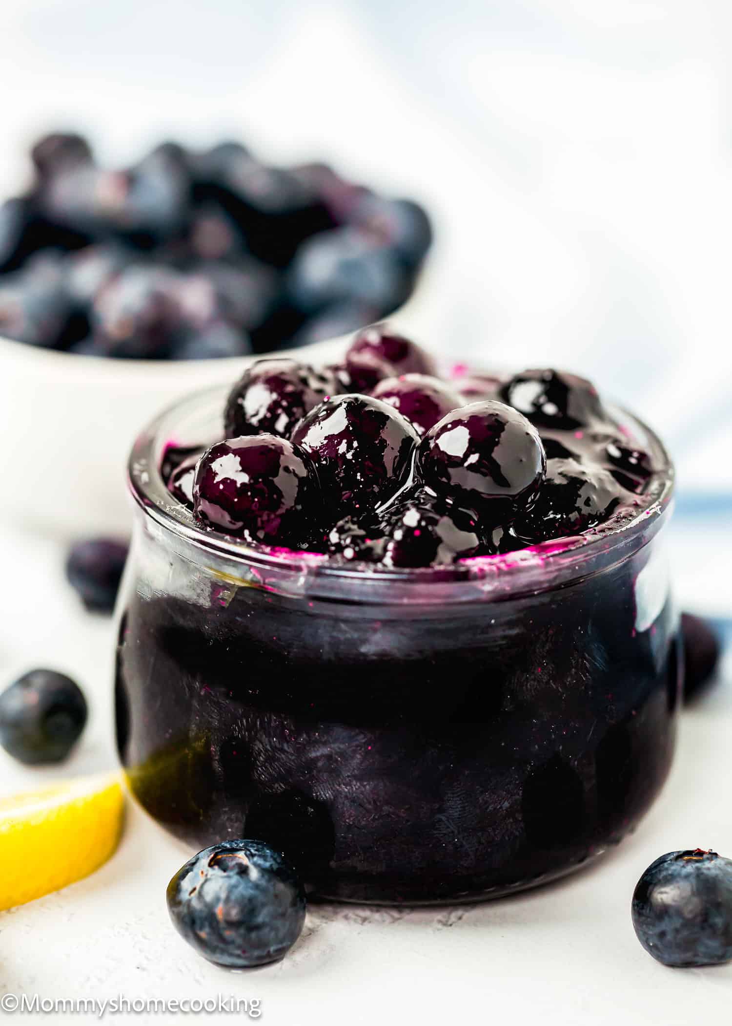 easy Blueberry syrup Topping in little glass jar over a white surface with more fresh blueberries and a lemon wedge on the side.