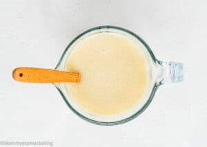Overhead view of a glass pitcher filled with a creamy, pale yellow liquid and a wooden spoon resting inside it on a white surface, ready to be poured into the batter for Eggless Zucchini Muffins.