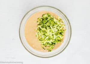 Shredded zucchini added to a bowl of tan batter, ready to be transformed into delicious Eggless Zucchini Muffins, viewed from above on a white surface.