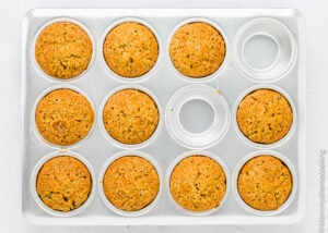 A 12-cup metal muffin tin with 10 baked, golden-brown Eggless Zucchini Muffins and 2 empty cups, viewed from above.