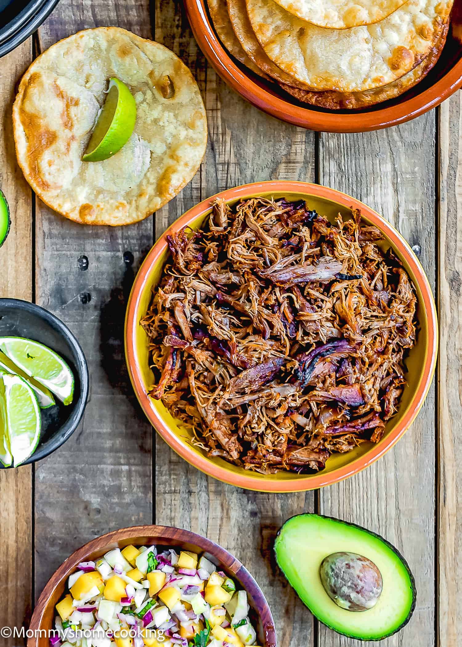Slow-Cooker Spicy Pork Carnitas in a bowl and tostadas, avocado and lime wedges on the sides over a wooden surface.