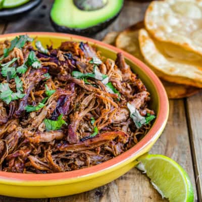 Slow-Cooker Spicy Pork Carnitas in a bowl with chopped cilantro on top and tostadas, avocado and lime wedges on the sides.