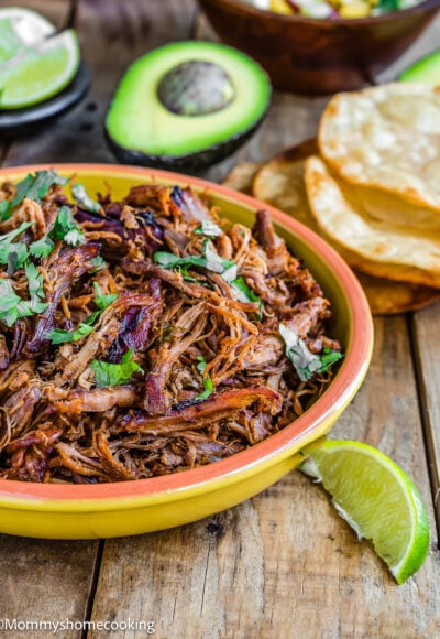 Slow-Cooker Spicy Pork Carnitas in a bowl with chopped cilantro on top and tostadas, avocado and lime wedges on the sides.