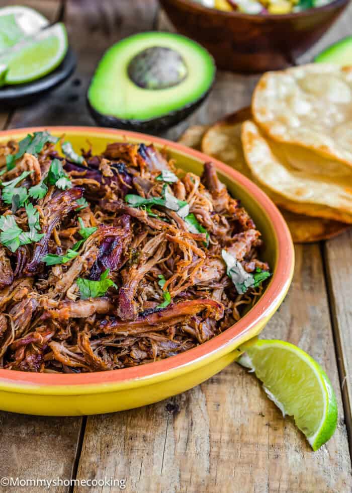 Slow-Cooker Spicy Pork Carnitas in a bowl with chopped cilantro on top and tostadas, avocado and lime wedges on the sides.