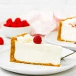a slice of Easy No Bake Cheesecake with a raspberry on top over a white dessert plate with a bowl of fresh raspberries and a kitchen towel in the background.