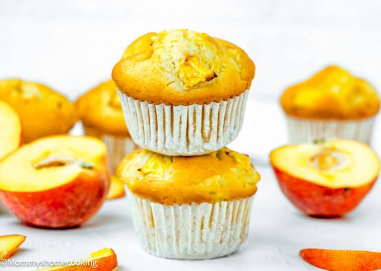 Two apple muffins stacked, surrounded by sliced apples and additional muffins, on a white surface with a bright background—perfect inspiration if you love wholesome bakes like Easy Peach Muffins (No Eggs - No Dairy).
