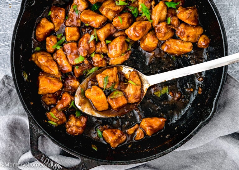 A skillet filled with Easy Sticky Bourbon Chicken pieces glazed to perfection and garnished with green herbs, with a metal spoon resting in the pan.