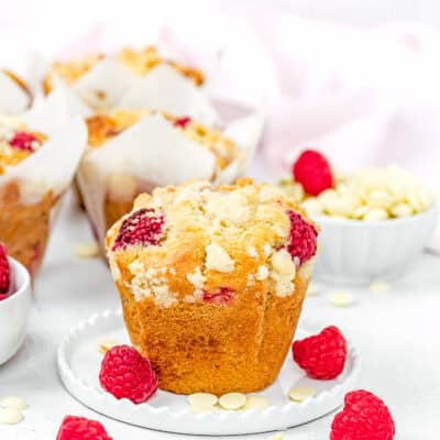 a Easy Vegan Raspberry Muffin on a little white plate with fresh raspberries and white chocolate around it with a more muffins in the background.