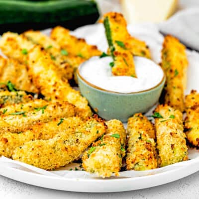 baked Eggless Zucchini Fries on a plate with Yogurt sauce and more zucchini, a kitchen towel, and a piece of Parmesan cheese in the background.