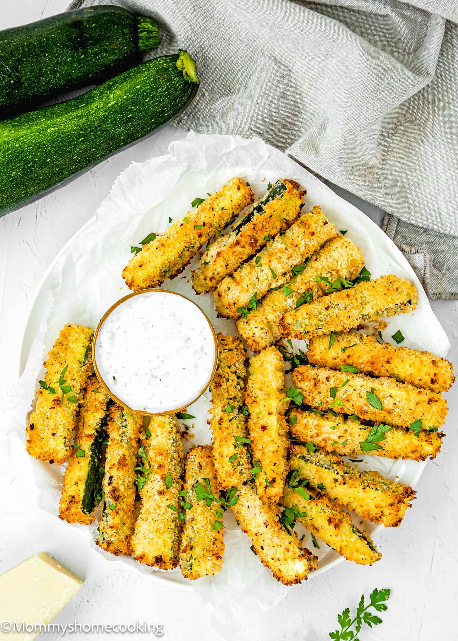 baked Eggless Zucchini Fries on a plate with Yogurt sauce and more zucchini, a kitchen towel, and a piece of Parmesan cheese around it.