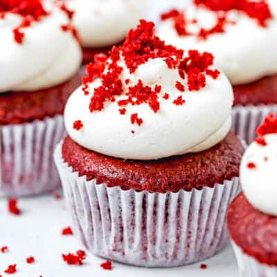 Egg-Free, Dairy-Free and Vegan Red Velvet Cupcake with frosting and red velvet crumbs on top with more cupcakes around.
