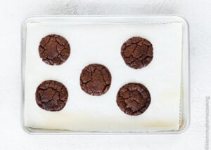 A baking tray lined with parchment paper holds five evenly spaced Best Eggless Brownie Cookies, fresh from the oven.