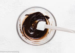 A glass bowl of melted chocolate being stirred with a white spatula on a white textured surface—perfect for making the Best Eggless Brownie Cookies.