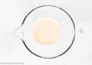 A glass mixing bowl containing a small amount of pale batter for Best Eggless Brownie Cookies sits on a white textured surface.