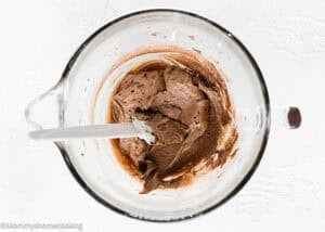 A glass mixing bowl containing rich chocolate batter for the Best Eggless Brownie Cookies, with a white spatula resting inside, viewed from above on a white surface.