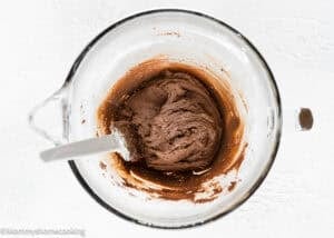 A glass mixing bowl with chocolate dough for the Best Eggless Brownie Cookies sits alongside a spatula on a white surface.
