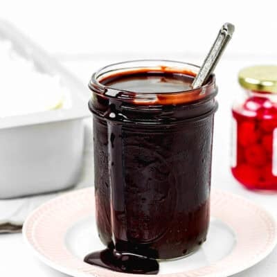 homemade Chocolate Hot Fudge Sauce in a jar over a plate, with vanilla ice cream and cherries in the background.