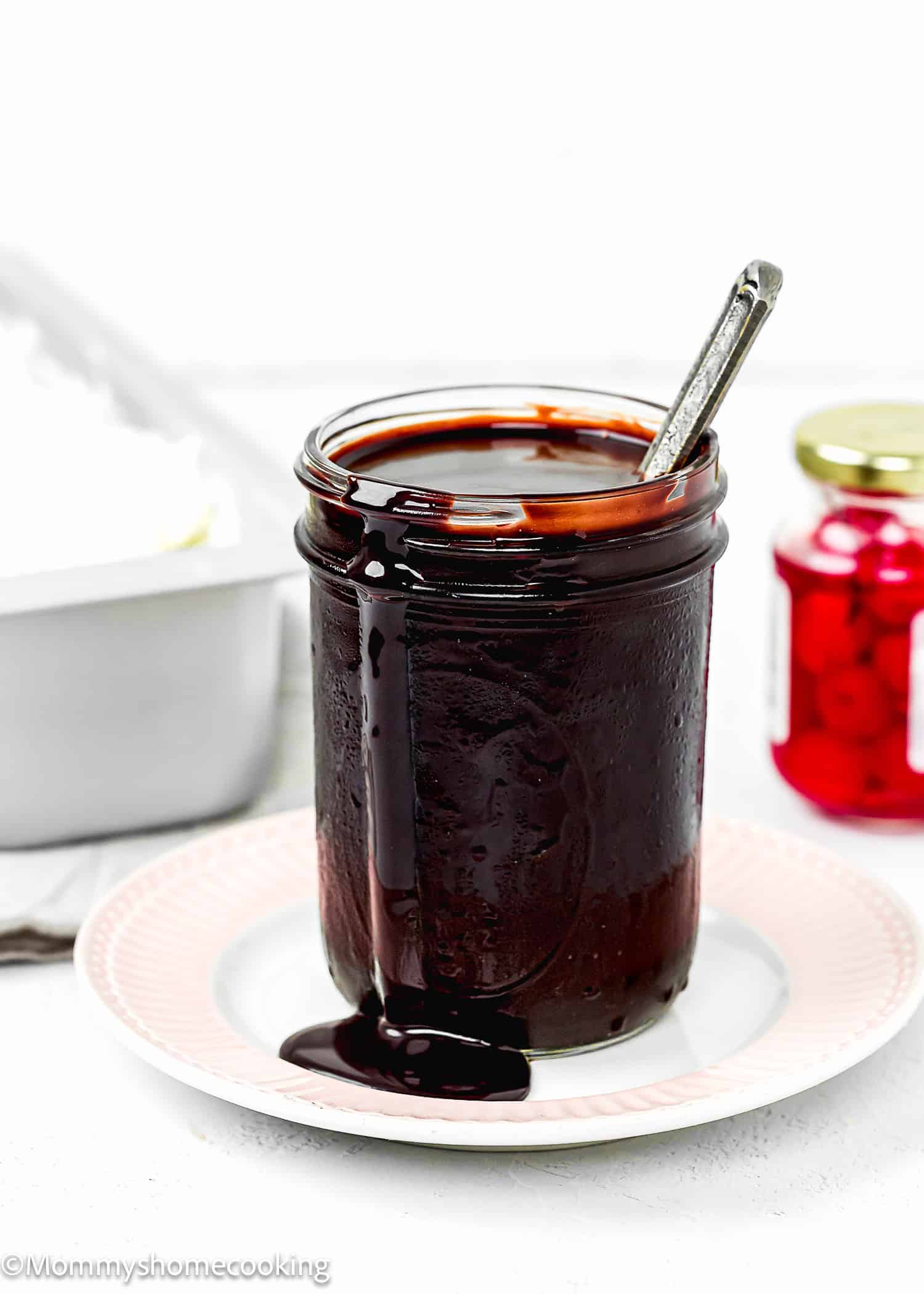 homemade Chocolate Hot Fudge Sauce in a jar over a plate, with vanilla ice cream and cherries in the background.