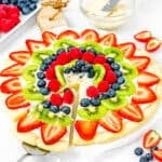 a cake server picking an Easy Egg-Free Fruit Pizza slice on a serving board over a white surface with more fresh fruit around it.