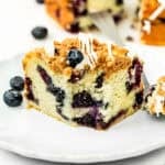 Eggless Easy Blueberry Crumb Cake slice on a plate with glaze and fresh blueberries.