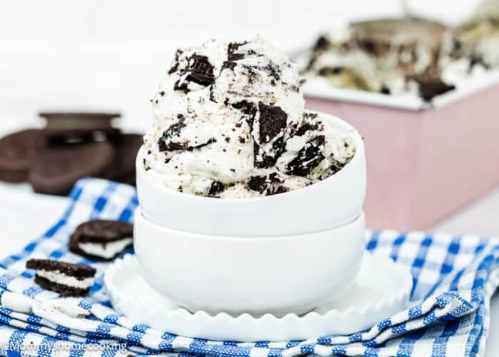 A bowl of Eggless Oreo Ice Cream (No-Churn Cookies & Cream) with cookie pieces, stacked on another empty bowl, rests on a blue checkered cloth with cookies in the background.