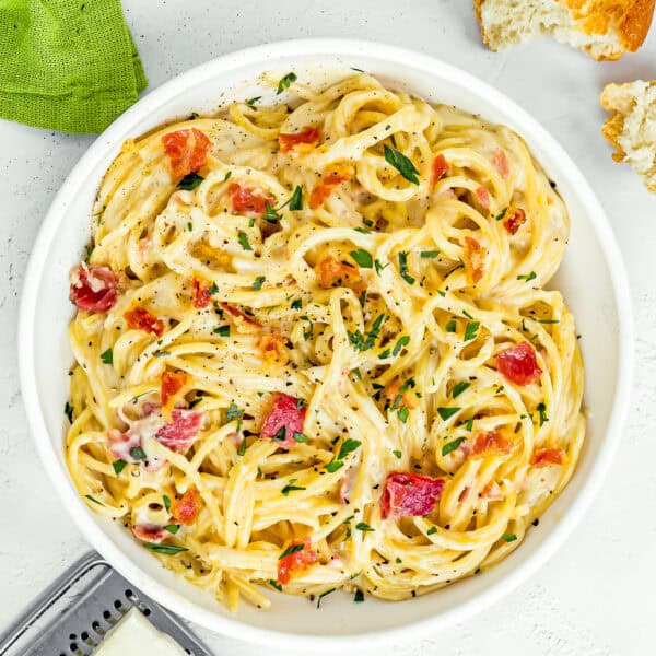 a bowl eggless Pasta Carbonara, garnished with chopped parsley with a green kitchen towel , cheese grater and bread on the sides.