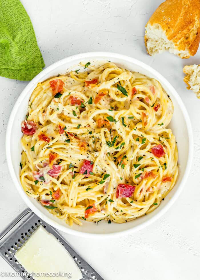 a bowl eggless Pasta Carbonara, garnished with chopped parsley with a green kitchen towel , cheese grater and bread on the sides.
