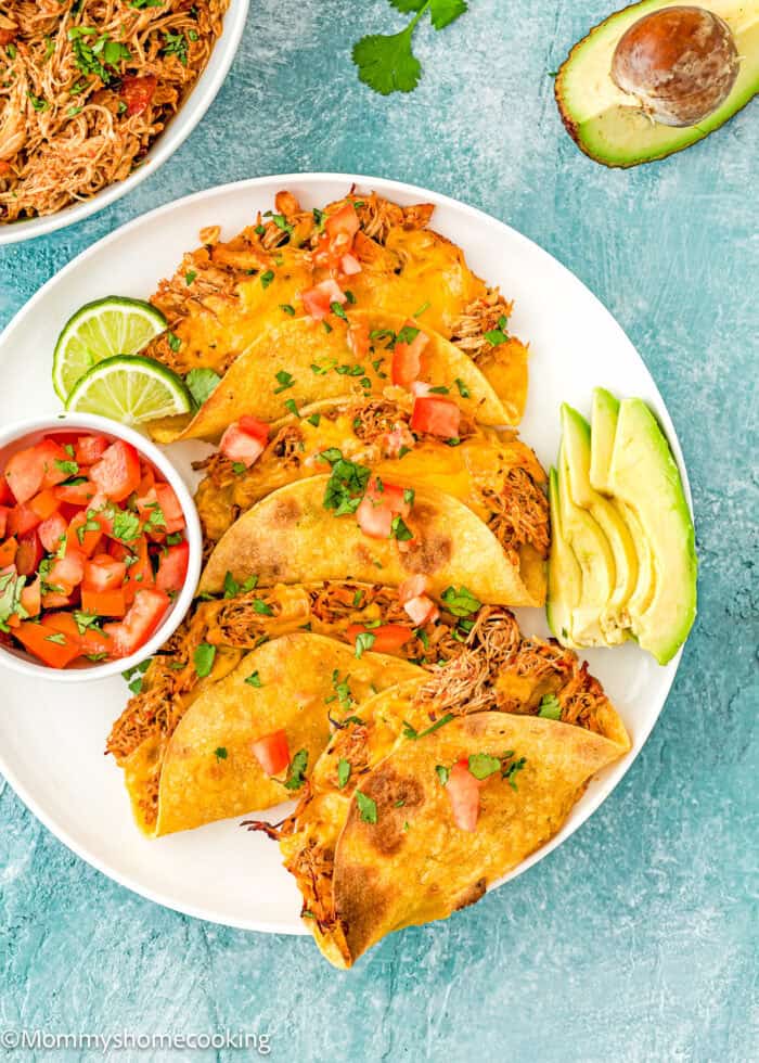 Oven-Baked Crispy Chicken Tacos on a plate with avocado and diced tomatoes over a blue surface.