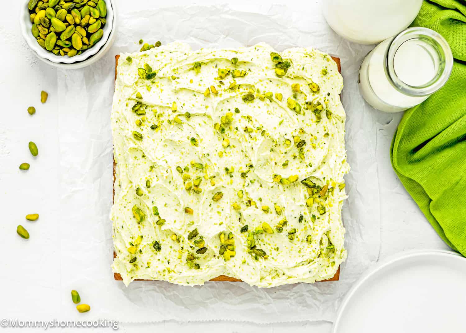 Egg-Free Easy Pistachio Cake with Pistachio buttercream over a white surface with more pistachios, plate and a glass of milk on the sides.