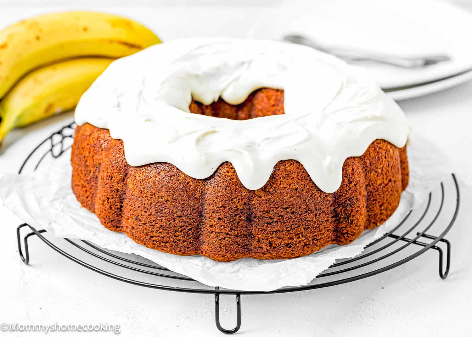 a whole Egg-free Brown Butter Banana Bundt Cake with glaze over a cooling rack with fresh bananas and plates on the background.