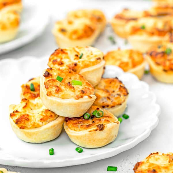 Eggless Easy Mini Quiches on a plate with more mini quiches on the sides over a white surface.