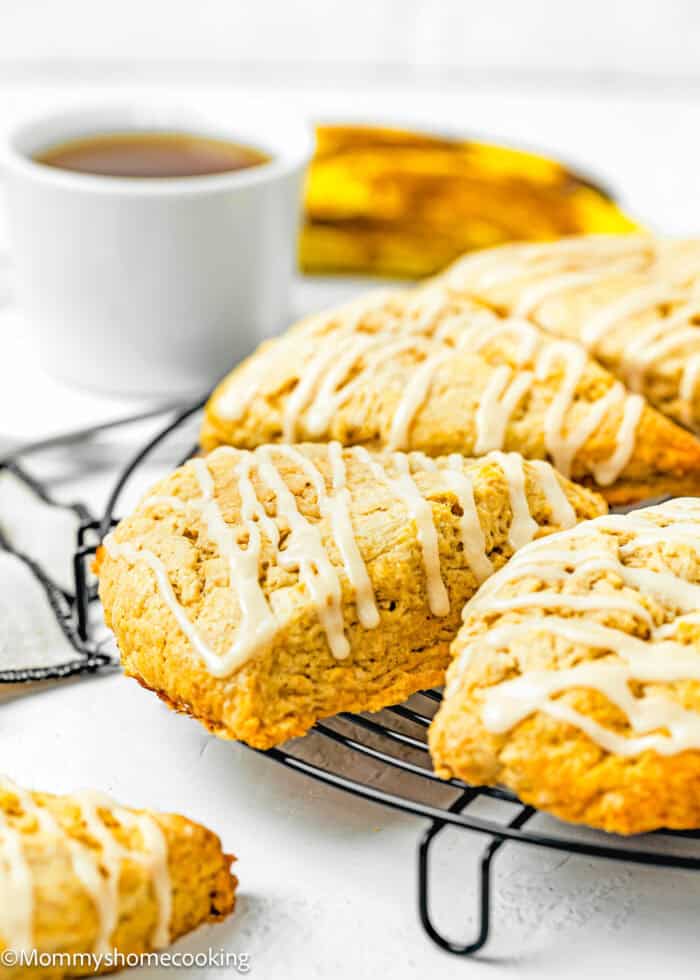 eggless Simple Banana Scones drizzled with glaze on a cooling rack over a white surface with a cup of tea on the background.