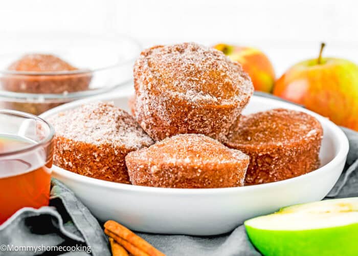 A white bowl filled with Easy Apple Cider Muffins (no eggs or dairy) is surrounded by apples, cinnamon sticks, and a cup of tea.