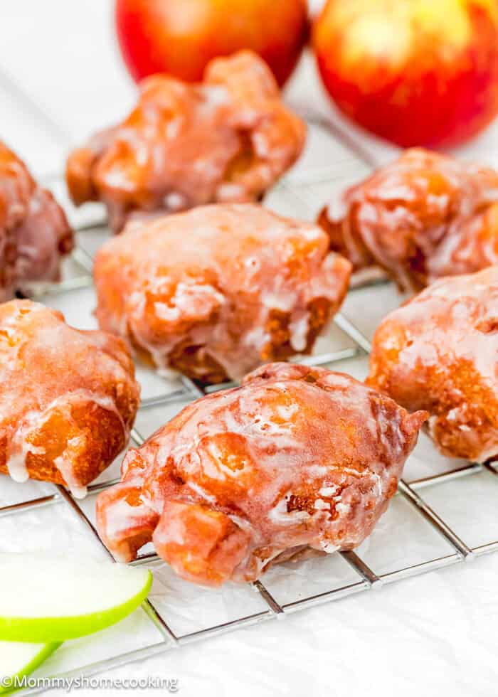 Eggless, easy apple fritters with a glazed coating are cooling on a wire rack. Apples are visible in the background.